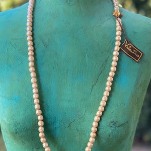 May include: A long, cream-colored pearl necklace with a gold clasp. The necklace is displayed on a mannequin with a teal-colored torso. The necklace has a small tag attached to it that reads "Valentino".