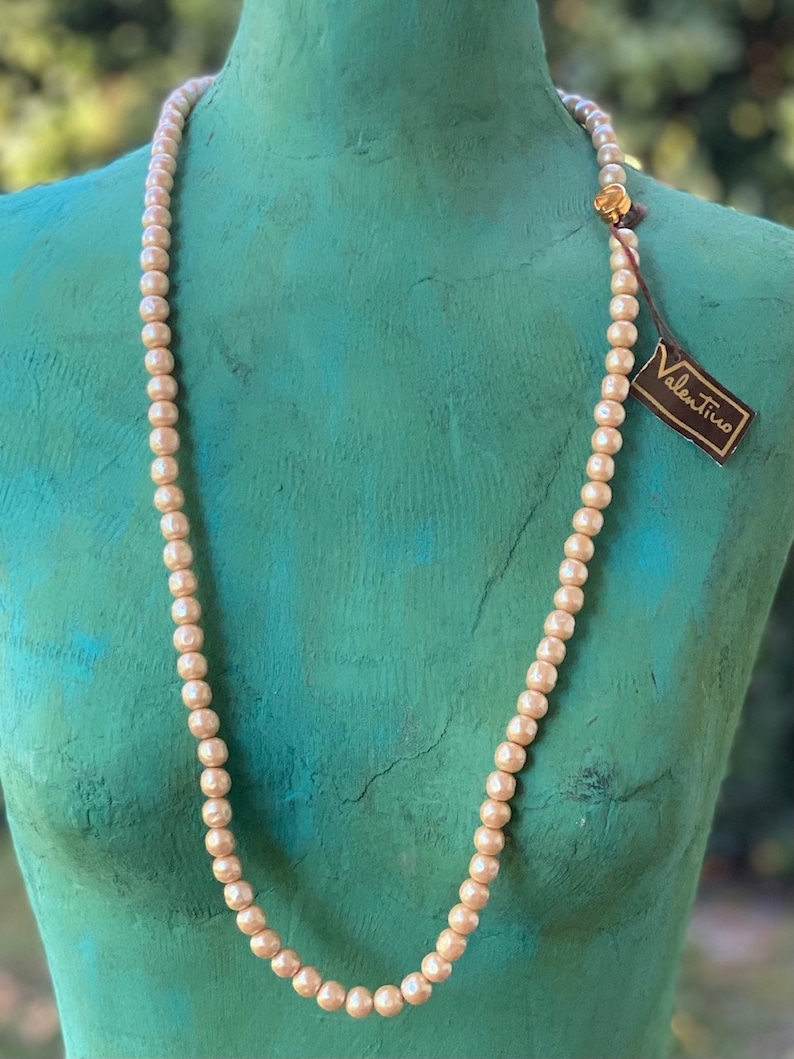 May include: A long, cream-colored pearl necklace with a gold clasp. The necklace is displayed on a mannequin with a teal-colored torso. The necklace has a small tag attached to it that reads "Valentino".