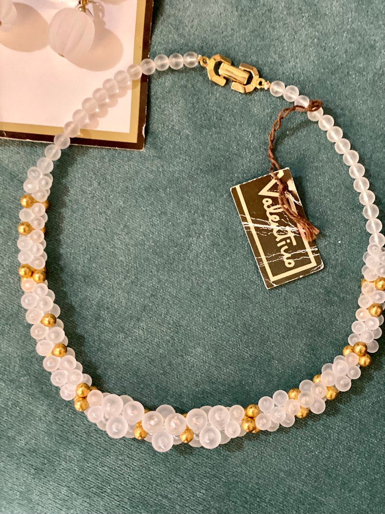 Puede incluir: Un collar de cuentas blancas y doradas con un cierre dorado. El collar est&aacute; hecho de cuentas peque&ntilde;as y redondas y tiene una etiqueta que dice "Valentino".