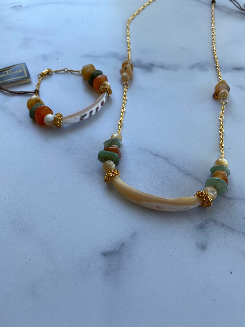 Puede incluir: Un collar y un brazalete a juego de cadena dorada con un colgante de concha y cuentas de piedras preciosas de colores.