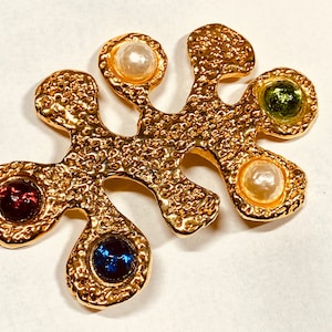 May include: Gold-toned brooch with a textured, abstract design. The brooch features five colorful stones: red, blue, green, white, and white.
