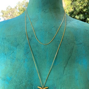 May include: A gold chain necklace with a gold dolphin tail pendant. The necklace is layered on a mannequin with a teal blue background.