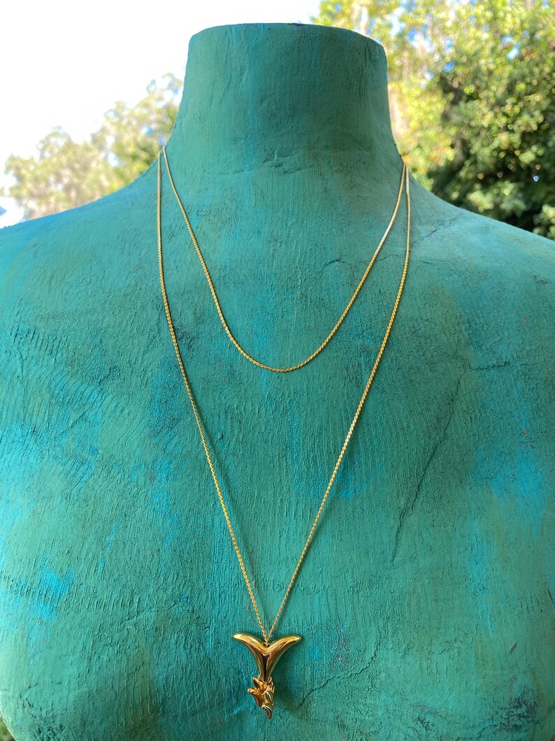 May include: A gold chain necklace with a gold dolphin tail pendant. The necklace is layered on a mannequin with a teal blue background.