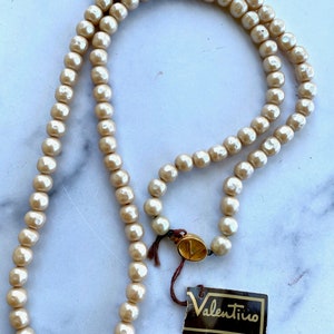 May include: A long, cream-colored pearl necklace with a gold clasp and a Valentino tag.