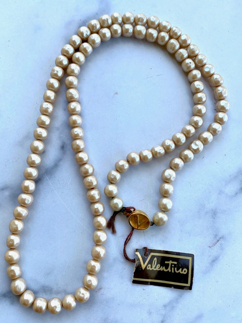 May include: A long, cream-colored pearl necklace with a gold clasp and a Valentino tag.