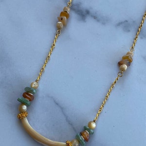 Puede incluir: Un collar de cadena dorada con un colgante de concha blanca curvado y cuentas verdes, marrones y blancas. El collar tiene dos racimos de cuentas en la cadena.