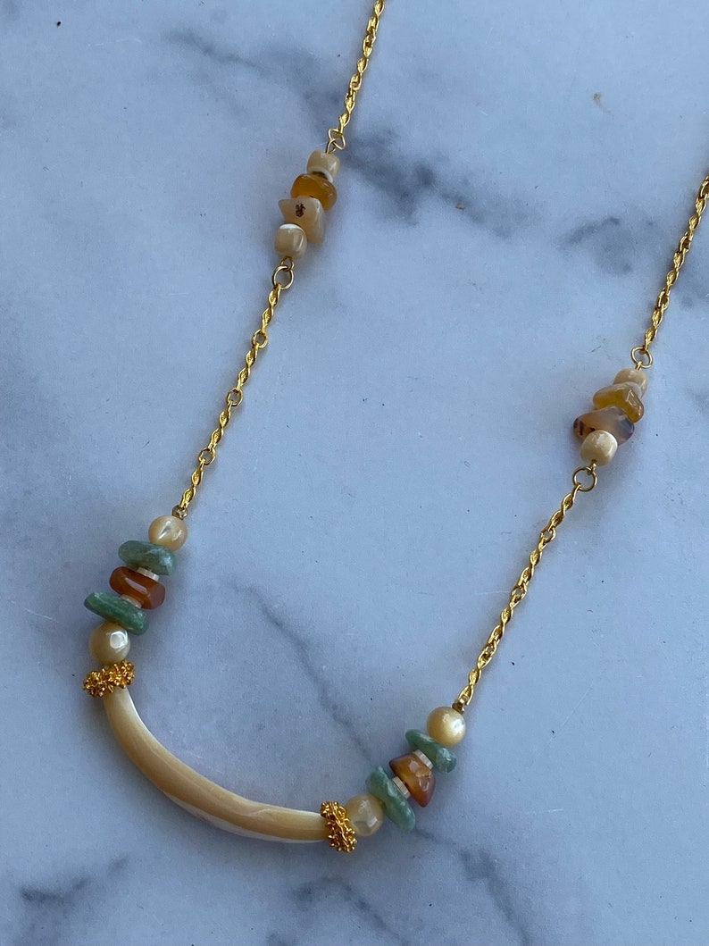 Puede incluir: Un collar de cadena dorada con un colgante de concha blanca curvado y cuentas verdes, marrones y blancas. El collar tiene dos racimos de cuentas en la cadena.