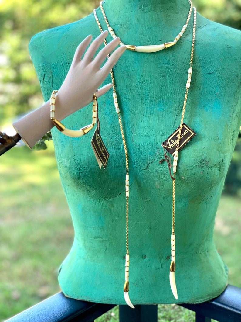 Puede incluir: Un conjunto de tres collares de cadena dorada con cuentas blancas de hueso y colgantes. Los collares se muestran en un maniqu&iacute; verde. La etiqueta en el collar dice "Valentino."