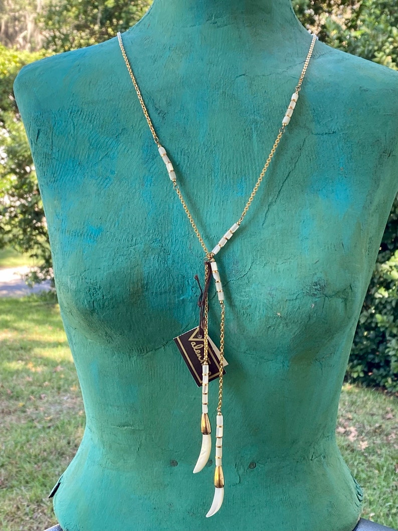 Puede incluir: Un collar de cadena dorada con cuentas de concha blanca y tres colgantes de concha blanca. El collar est&aacute; en un maniqu&iacute; con un torso verde azulado.