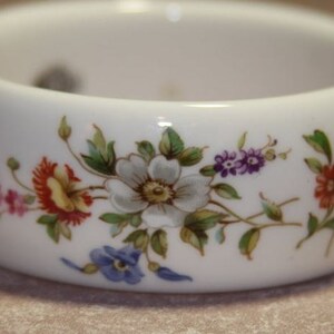 Peut inclure: Un anneau de serviette en c&eacute;ramique blanche avec un motif floral compos&eacute; de fleurs roses, rouges, blanches, jaunes et violettes. Les fleurs sont peintes sur l'anneau et ont des feuilles vertes.