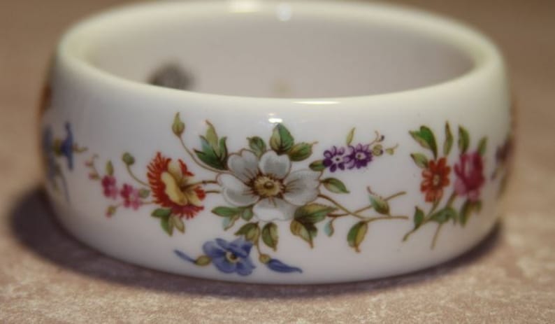 Peut inclure: Un anneau de serviette en c&eacute;ramique blanche avec un motif floral compos&eacute; de fleurs roses, rouges, blanches, jaunes et violettes. Les fleurs sont peintes sur l'anneau et ont des feuilles vertes.