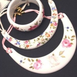 Peut inclure: Un ensemble de trois colliers en c&eacute;ramique blanche avec des motifs floraux. Les colliers sont en forme de cercle, de croissant de lune et de demi-lune. Les fleurs sont roses, jaunes et bleues.