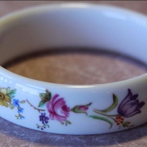 May include: White porcelain bangle bracelet with a floral design. The flowers are pink, yellow, purple, and blue.