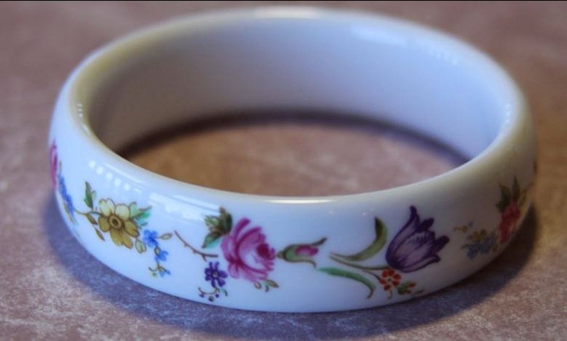 May include: White porcelain bangle bracelet with a floral design. The flowers are pink, yellow, purple, and blue.