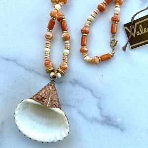 May include: A long necklace with orange and white beads and a large, white seashell pendant with gold accents. The necklace has a gold clasp and a tag that says "Valentino".
