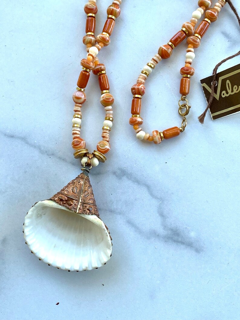 May include: A long necklace with orange and white beads and a large, white seashell pendant with gold accents. The necklace has a gold clasp and a tag that says "Valentino".