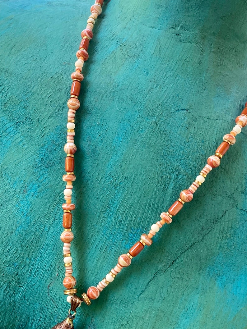 May include: A long necklace made of brown, white, and pink beads with a gold pendant at the bottom.