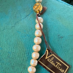 May include: A cream-colored pearl necklace with a gold clasp and a brown tag that reads "Valentino".
