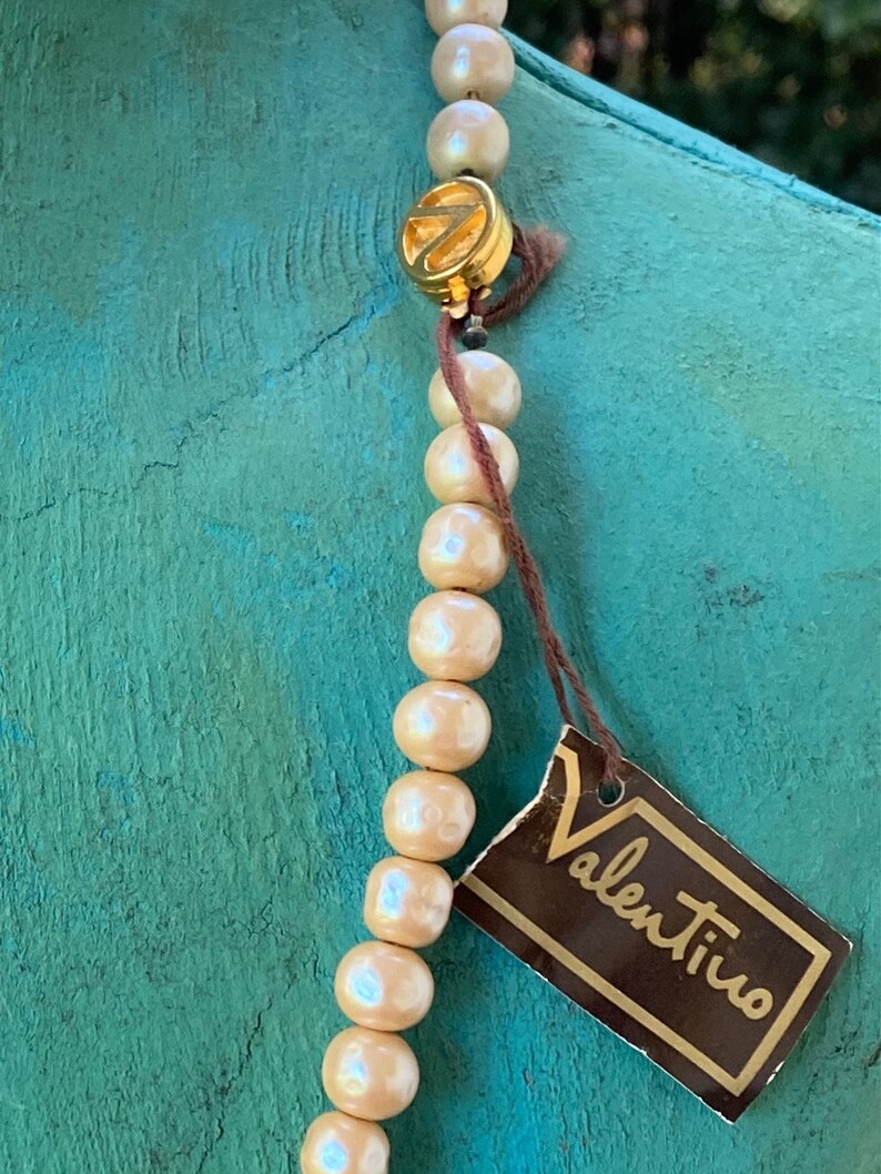 May include: A cream-colored pearl necklace with a gold clasp and a brown tag that reads "Valentino".