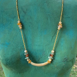 Puede incluir: Un collar de cadena dorada con un colgante blanco curvo y cuentas multicolores. Las cuentas son una mezcla de marr&oacute;n, beige y verde.