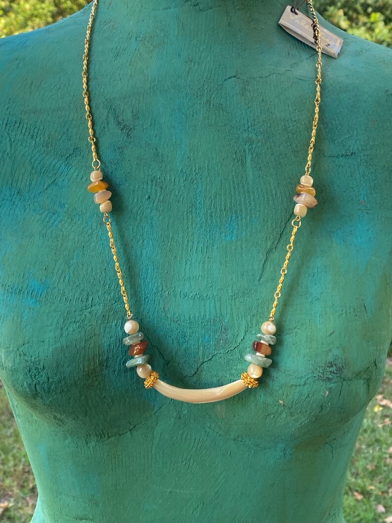 Puede incluir: Un collar de cadena dorada con un colgante blanco curvo y cuentas multicolores. Las cuentas son una mezcla de marr&oacute;n, beige y verde.