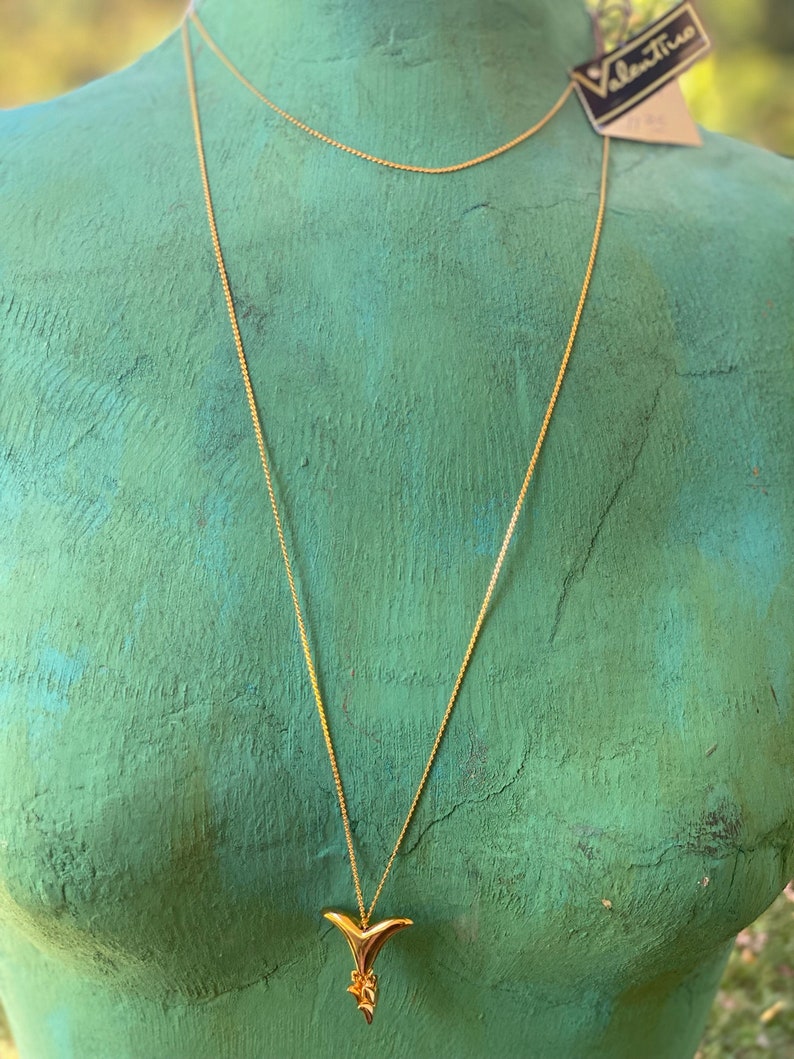 May include: A gold chain necklace with a gold pendant shaped like a stylized bird. The necklace is on a mannequin with a green background.