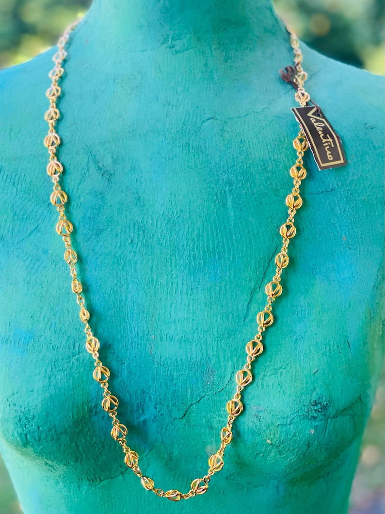May include: A gold chain necklace with a delicate, openwork design. The chain is made of small, linked circles with a cutout pattern. The necklace is displayed on a turquoise mannequin.