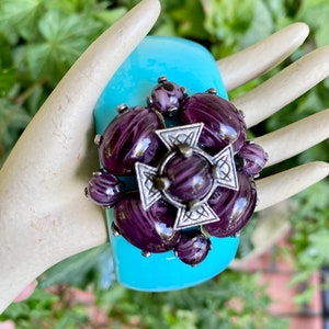 May include: A turquoise plastic bangle bracelet with a large purple and silver brooch. The brooch is a cluster of purple cabochons with a silver cross in the center.