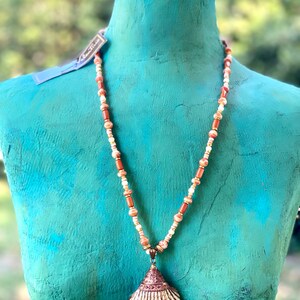 May include: A necklace with a large seashell pendant. The necklace is made of brown and white beads. The seashell is a light brown color with a white interior.