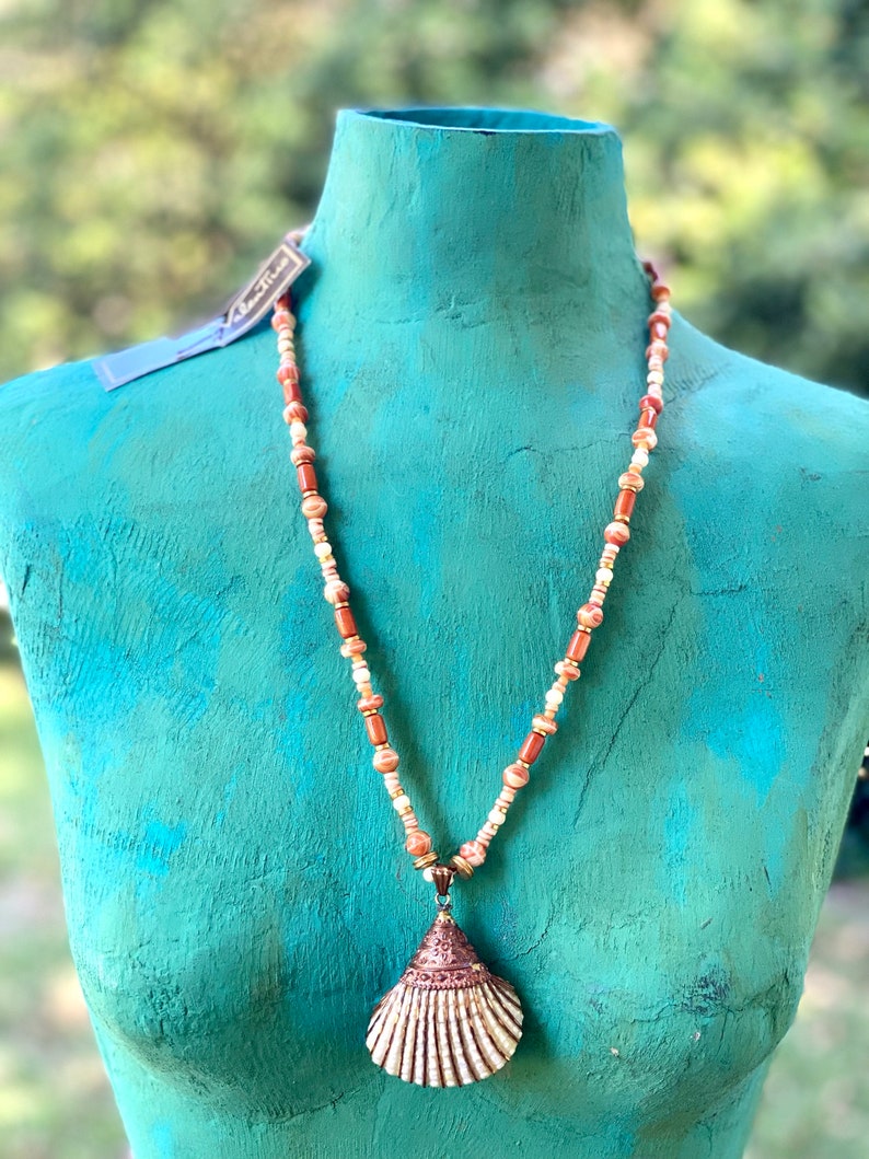 May include: A necklace with a large seashell pendant. The necklace is made of brown and white beads. The seashell is a light brown color with a white interior.