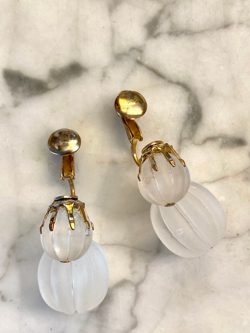 Puede incluir: Un par de pendientes de clip de tono dorado con colgantes de vidrio blanco esmerilado en forma de l&aacute;grima.