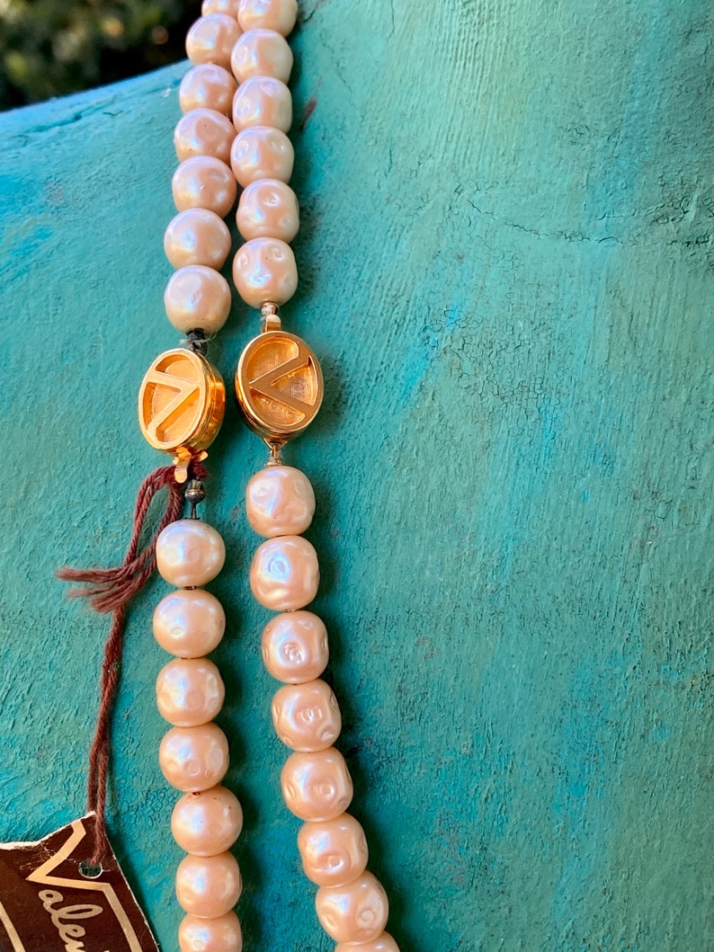 May include: A double strand pearl necklace with gold clasps. The clasps are round and have a letter 'V' design. The pearls are cream colored and have a slight iridescent sheen.