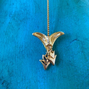 May include: Gold tone pendant necklace with a fishtail charm and two smaller arrow charms. The necklace is hanging against a turquoise background.