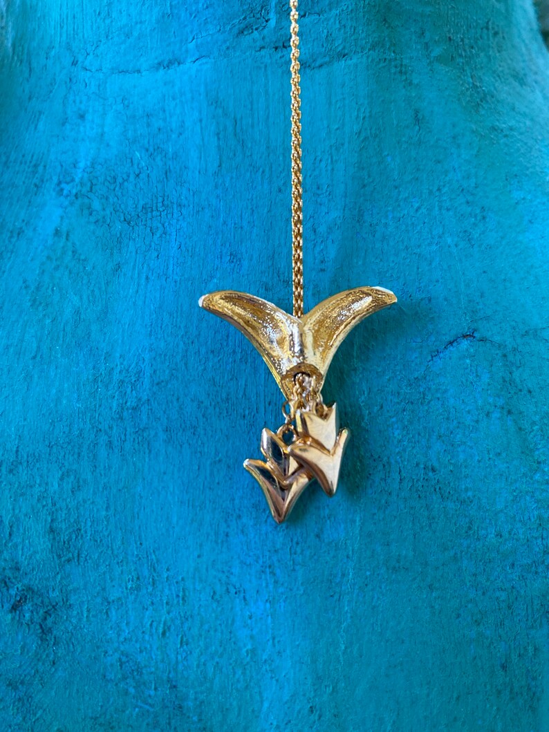May include: Gold tone pendant necklace with a fishtail charm and two smaller arrow charms. The necklace is hanging against a turquoise background.