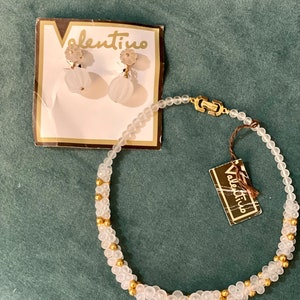 Puede incluir: Un collar de cuentas blancas con detalles dorados y pendientes a juego. El collar y los pendientes est&aacute;n etiquetados como "Valentino".