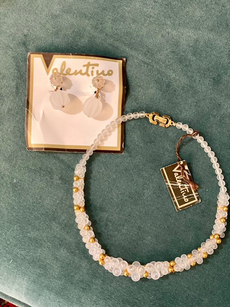 Puede incluir: Un collar de cuentas blancas con detalles dorados y pendientes a juego. El collar y los pendientes est&aacute;n etiquetados como "Valentino".