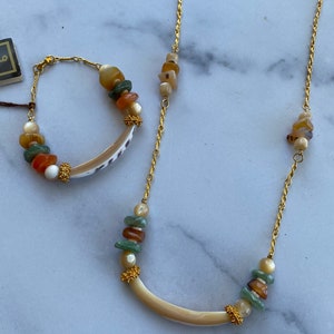 Puede incluir: Un collar y un brazalete de cadena dorada con un colgante de concha curvado y cuentas de piedras multicolores.