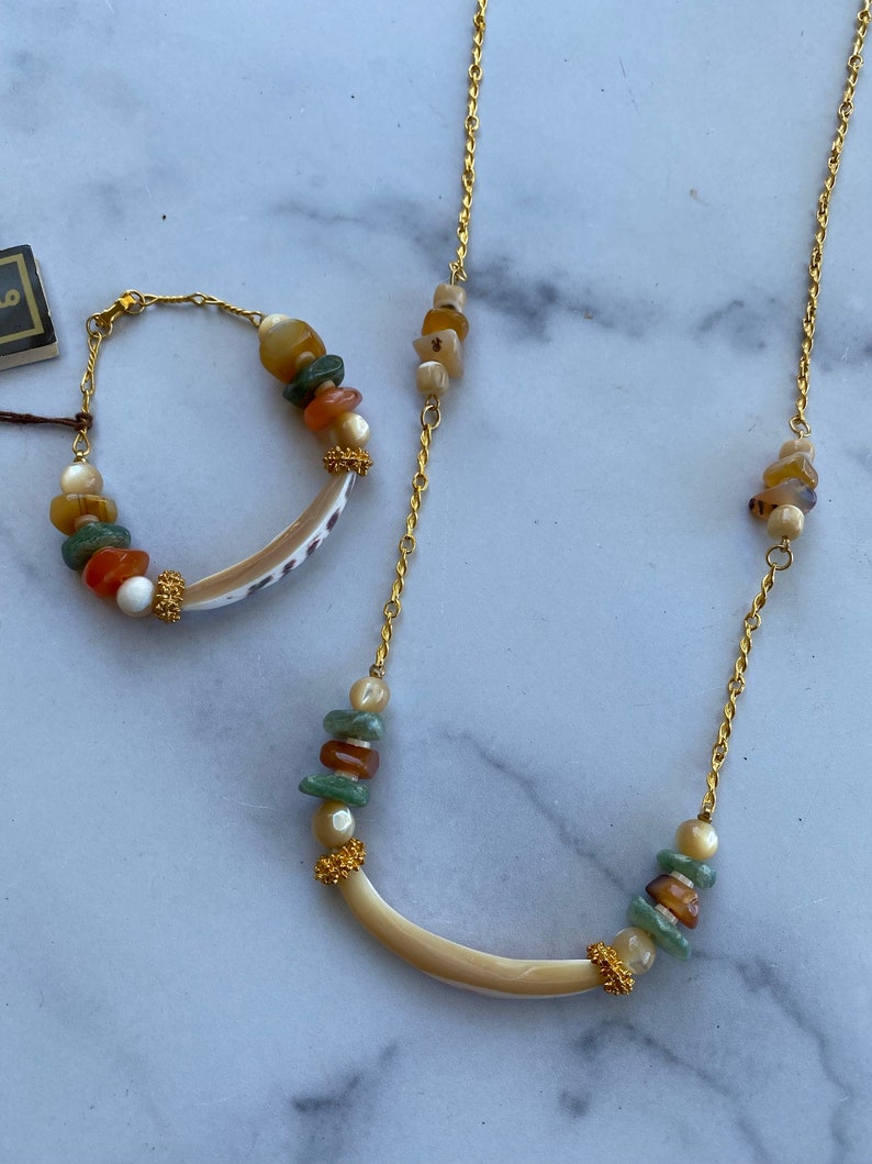 Puede incluir: Un collar y un brazalete de cadena dorada con un colgante de concha curvado y cuentas de piedras multicolores.