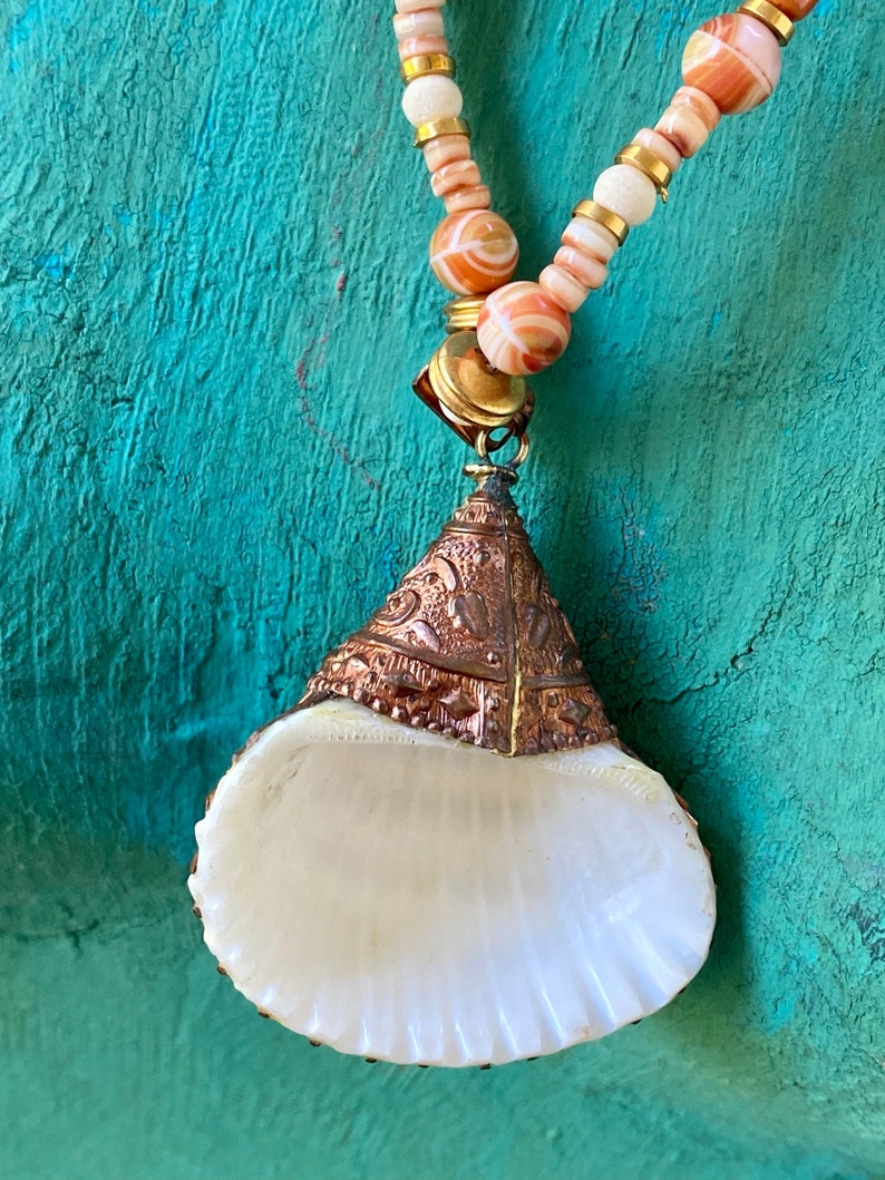 May include: A white seashell pendant necklace with a copper and gold-toned metal top. The necklace is made with white and orange striped beads and gold-toned beads.
