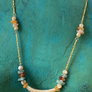 Puede incluir: Un collar de cadena dorada con un colgante curvo de concha blanca. El collar presenta cuentas multicolores de forma irregular.