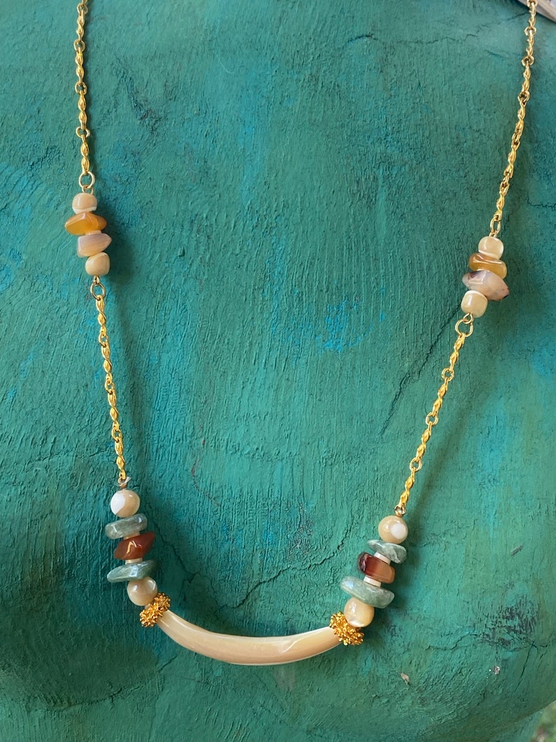 Puede incluir: Un collar de cadena dorada con un colgante curvo de concha blanca. El collar presenta cuentas multicolores de forma irregular.