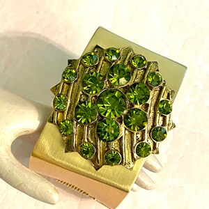 May include: A gold metal cuff bracelet with a large green rhinestone cluster. The rhinestones are set in a circular pattern and have a faceted design.