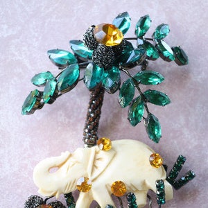 May include: A white elephant figurine with green and gold rhinestones, standing under a green rhinestone tree with a black metal base.