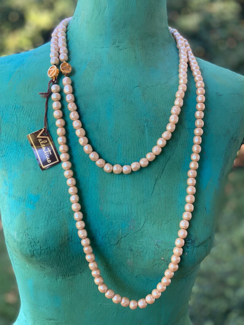 May include: A long, double-strand pearl necklace with gold clasps. The necklace is displayed on a mannequin with a teal-colored torso. The necklace has a tag that reads "Valentina".