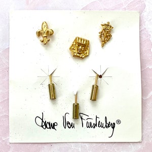 May include: A white card with gold-colored metal pins and the text "Diane von Furstenberg" in black ink. The pins feature a fleur-de-lis, a crown, and a face.