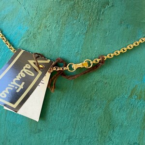 Puede incluir: Un collar de cadena dorada con una cuerda marr&oacute;n atada a un cierre. El collar tiene una etiqueta adjunta que dice "Glamour" en letras negras.