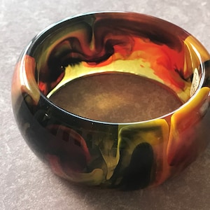 May include: A black, yellow, and orange swirl patterned bangle bracelet. The bangle is made of plastic and has a glossy finish.