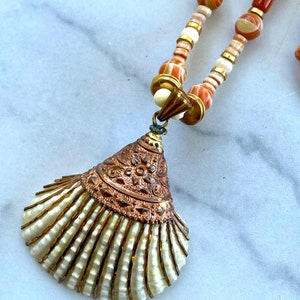 May include: A long necklace with a shell pendant. The necklace is made of brown and white striped beads with gold accents. The shell pendant is white with gold accents and a detailed design.