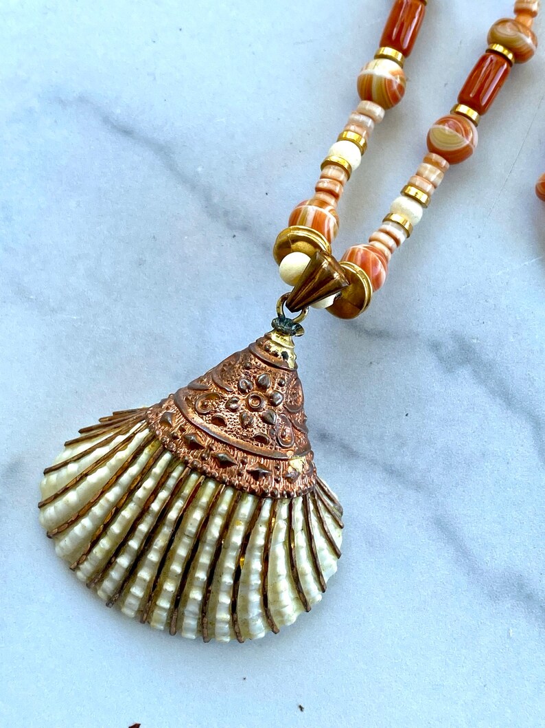 May include: A long necklace with a shell pendant. The necklace is made of brown and white striped beads with gold accents. The shell pendant is white with gold accents and a detailed design.