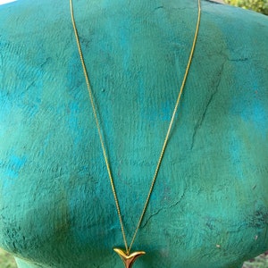 May include: A gold chain necklace with a gold shark fin pendant. The necklace is on a mannequin with a teal blue torso. The tag on the necklace reads "alexine".